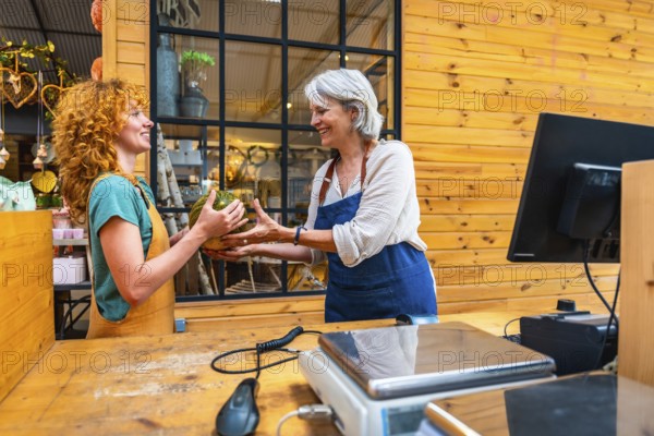 Two women joyfully exchanging a pumpkin in a vibrant flower shop, with a scale and computer prominently displayed on the counter