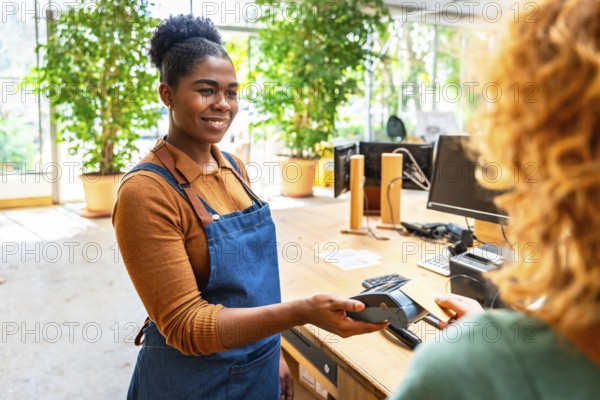 Smiling cashier holding pos terminal while customer paying with smartphone using nfc technology in a plant store