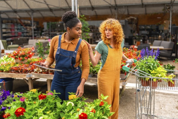 Two women working in a garden center are discussing flowers while using a digital tablet and pushing a shopping cart