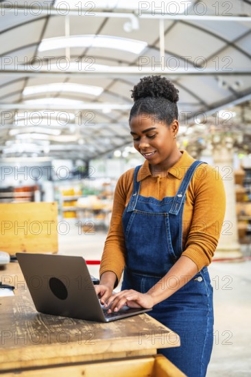 Young woman wearing denim overalls typing on laptop in plant nursery, managing inventory and online orders