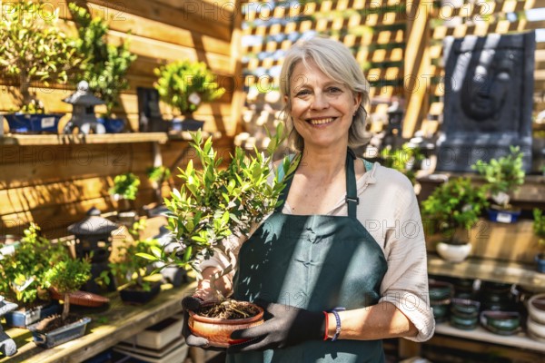 Senior gardener woman smiling while holding a small bonsai tree in a vibrant shop, showcasing her passion for nurturing plants