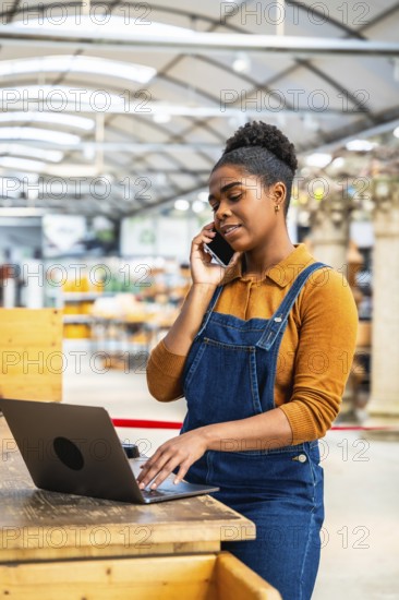Confident black female employee managing orders and assisting customers in a garden center, using laptop and smartphone simultaneously