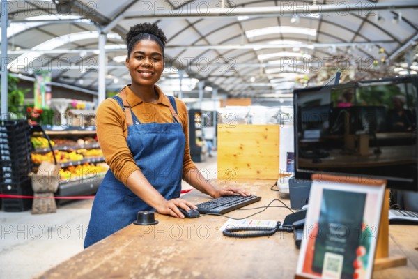 Young female cashier smiling and working at cash register in supermarket with fresh produce in background
