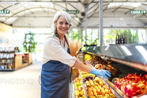 Smiling greengrocer wearing protective gloves, carefully arranging a vibrant display of fresh fruit in a bustling supermarket environment