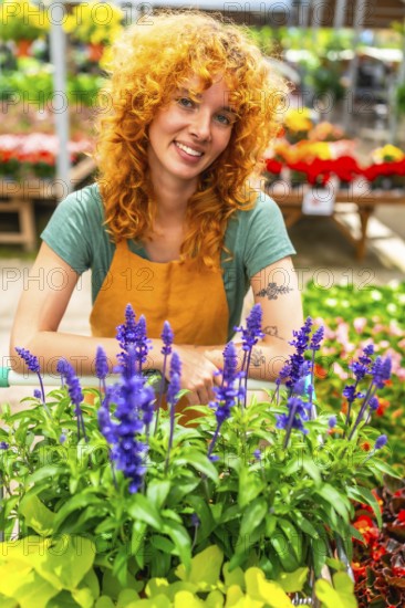 Smiling redhead gardener standing behind a cart overflowing with vibrant purple flowers, surrounded by the lush greenery of a greenhouse