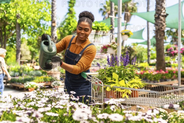 Happy young woman working in a garden center watering plants with a watering can while pushing a shopping cart