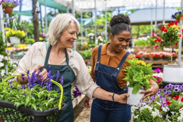 Two happy multiethnic female garden center workers carrying plants and flowers in a greenhouse