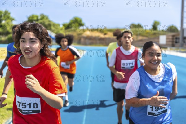 Teenage track and field athletes running on a blue track during a competition, wearing bibs with their names and numbers