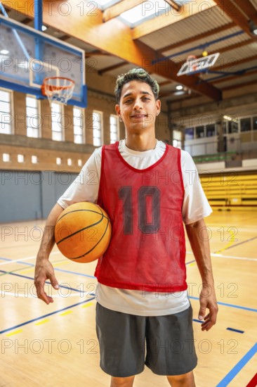 Energetic young basketball player holding a ball on an indoor court, wearing a red jersey, ready for practice or a game in a sports facility