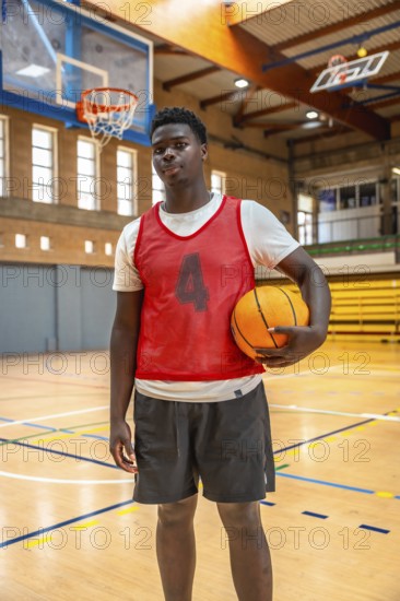 Young male basketball player holding a ball while standing confidently on a polished wooden court inside a vibrant gymnasium