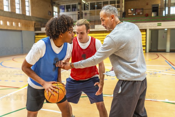 Basketball coach holding clipboard explaining game strategy to two players practicing during training in a gym