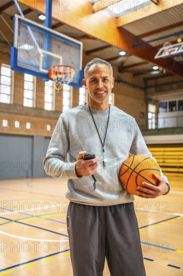 Mature basketball coach holding a ball and stopwatch, smiling confidently in an empty gymnasium, embodying passion for the sport