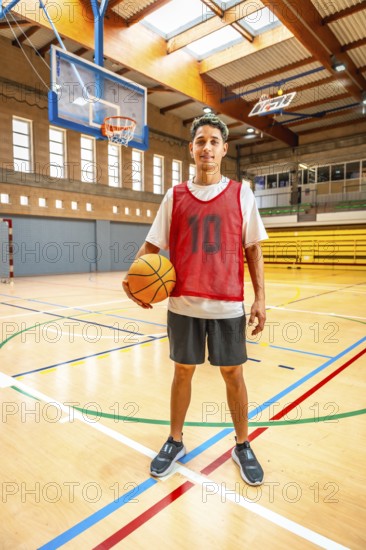 Young basketball player standing confidently on the court, holding a basketball, ready for the game in a brightly lit gymnasium