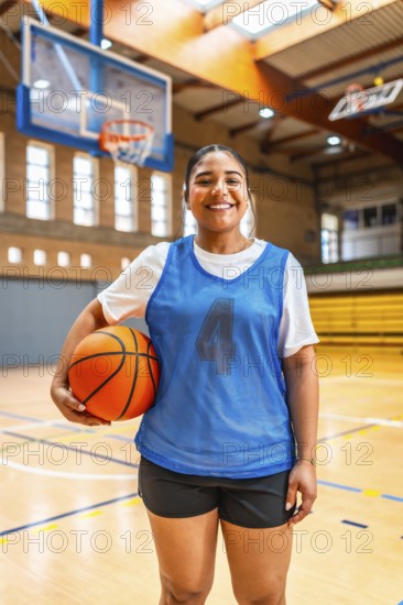 Young female athlete wearing blue jersey and black shorts, holding basketball and smiling in an indoor court