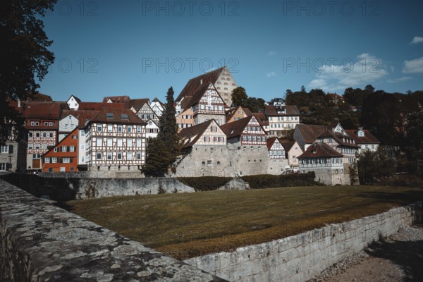 Medieval half-timbered houses and a castle under a blue sky next to a green meadow and trees in the town of Schwäbisch Hall, Baden-Württemberg, Germany