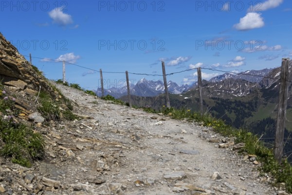 Rocky path with wooden fence, winding through a rugged, alpine mountain landscape under a blue sky