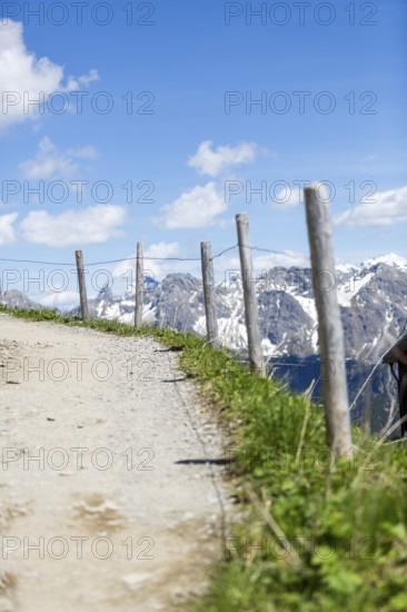 Narrow path with wooden posts at the edge, leads through an alpine landscape with snow-covered mountains