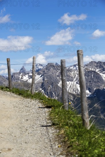 A path lined with wooden fences leads through an alpine landscape with imposing mountain peaks