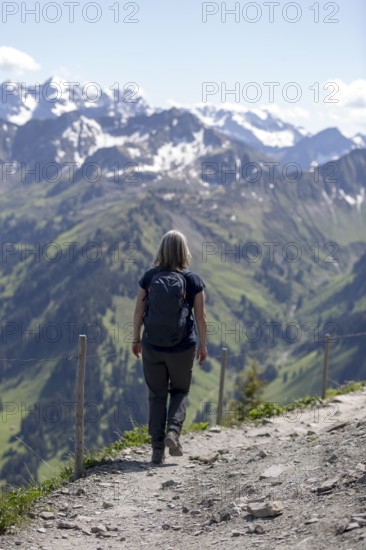 A hiker on a mountain path over green slopes with a view of the vast, snow-covered mountain landscape of the Alps