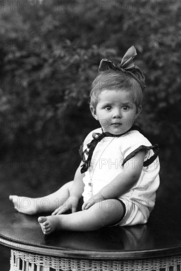 Little girl sitting on a table with a bow in her hair, portrait of a baby with a bow in children's clothes sitting on a table, historical photo