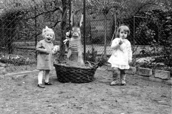Easter bunny basket girl, 1920, Two small children standing in a garden next to a large basket with a bunny, the surroundings are cheerful, Historical photo