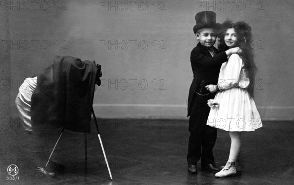 Children wedding couple photographer, 1920, Two children in formal wear smiling and posing in front of an antique camera as if they are dancing, Historical photo