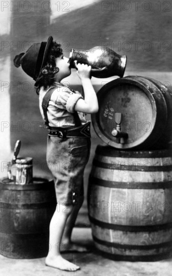 Child drinking wine, 1910s, A boy in traditional dress drinks from a large jug next to beer barrels, Historical photo