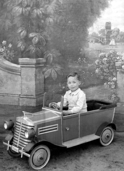 Little boy in car, 1930s, A boy sits in a toy car in front of a picturesque background, Historical photo
