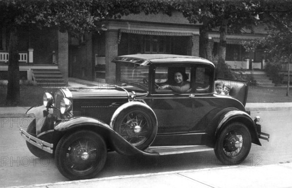 Woman in vintage car children, 1930s, A vintage car with passengers drives along a street with classic houses, Historical photo