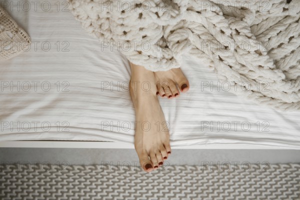 Bare feet with red nail polish rest on crisp white sheets, partially covered by a soft, knitted blanket. The cozy atmosphere invites relaxation and comfort in a serene bedroom setting