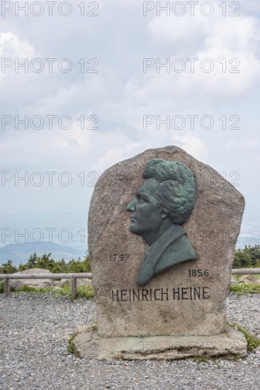 Heinrich Heine memorial, Heine memorial on the summit of the Brocken, summit plateau, granite stone with Heine plaque, portrait of the poet on bronze, inscription, year of birth, year of death, rock, forest of spruces, hazy, veiled view of the north-eastern Harz foreland, view with cloudy sky, Brocken, Harz National Park, Harz, Saxony-Anhalt, Germany