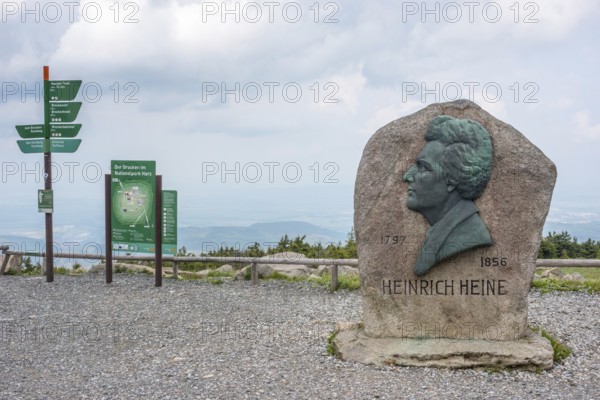 Heinrich Heine memorial, Heine memorial on the summit of the Brocken, summit plateau, granite stone with Heine plaque, portrait of the poet on bronze, inscription, year of birth, year of death, hiking map, map and signpost, hazy, veiled view of the north-eastern Harz foreland, view with cloudy sky, Brocken, Harz National Park, Harz, Saxony-Anhalt, Germany