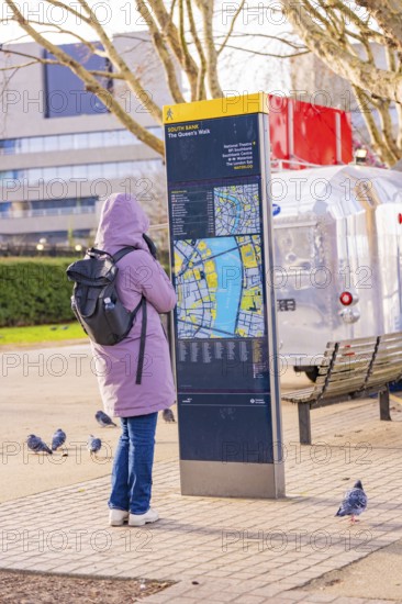 Woman in warm clothes looking at a map at a city map kiosk, pigeons on the ground, London, United Kingdom, World