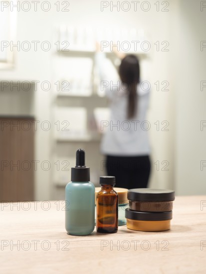 Skincare and cosmetic product bottles are on a table with a blurred scientist arranging items in the background