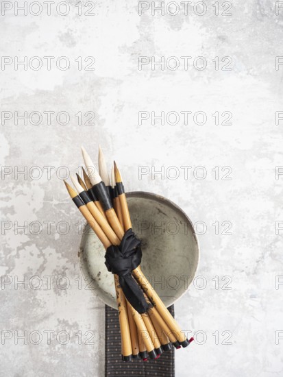 Multiple bamboo paint brushes tied with a black ribbon are resting on a ceramic bowl on a textured gray surface