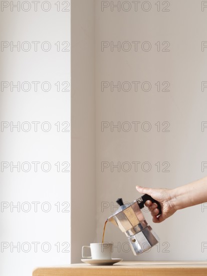 Hand pouring coffee from a moka pot into a cup on a wooden table against a minimalist white background