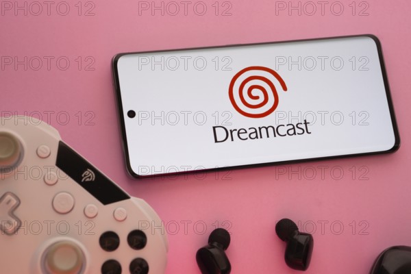 Dhaka, Bangladesh- 08 Apr 2025: A smartphone displaying the Dreamcast logo next to a gaming controller and earbuds on a pink surface