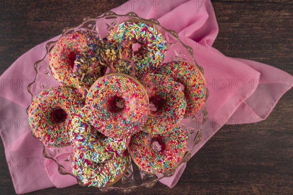 Donuts with colorful sprinkles on glass plate and pink fabric on wooden table