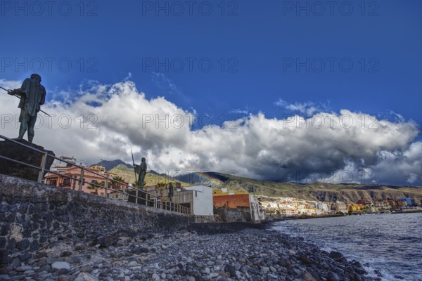 Coastline with statues and houses under a cloudy sky along a rocky shore, Candelaria Tenerife