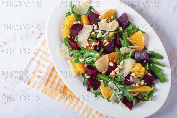 A salad made with chunks of beet, orange slices, and arugula. It is topped with cheese shavings and pine nuts, placed on a white plate with a striped napkin beside it