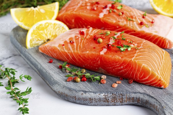 Two pieces of salmon fillet sit on a wooden cutting board. They are topped with herbs and pink peppercorns. Slices of lemon and lime surround the fish, creating a vibrant setting for cooking