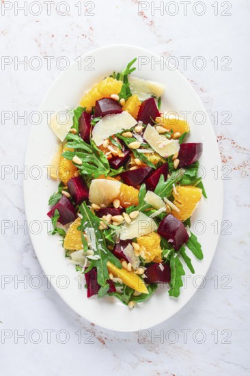A salad made with chunks of beet, orange slices, and arugula. It is topped with cheese shavings and pine nuts, placed on a white plate with a striped napkin beside it