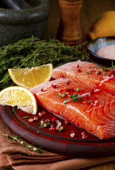 Two salmon fillets rest on a wooden platter. The fillets are garnished with herbs and spices. Lemon and lime slices are nearby. This setup is ready for preparation in a kitchen