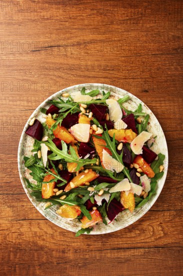 A bowl of salad contains beetroot, orange slices, pine nuts, and leaves. It is placed on a wooden table. The meal is healthy and looks colorful