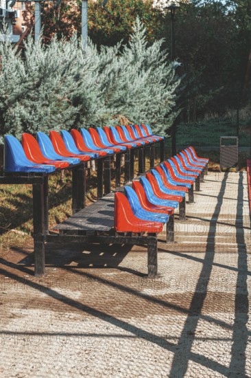 Rows of red and blue stadium seats are neatly arranged in a park. Bright sunlight illuminates the seating area, creating contrasting shadows on the ground