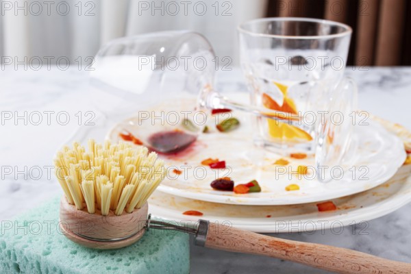 Dishes are stacked on a kitchen counter with food remnants after a meal. A cleaning brush and sponge are ready for washing the plates and glasses