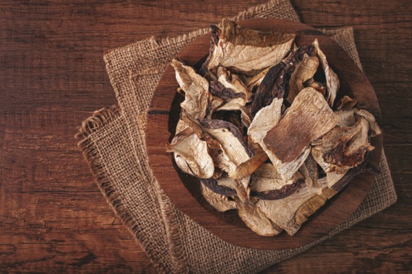 A collection of dried mushrooms fills a wooden bowl placed on a cloth surface. The mushrooms are cut into thin slices, ideal for adding to various dishes
