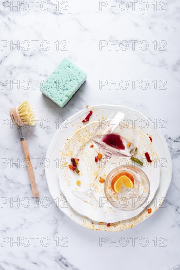Dishes are stacked on a kitchen counter with food remnants after a meal. A cleaning brush and sponge are ready for washing the plates and glasses