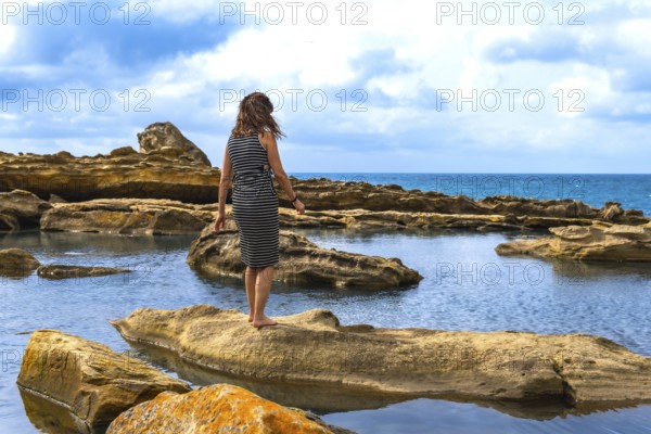 Woman standing barefoot on textured sandstone by tidal pools, facing the vast sea under a cloudy sky, serene and contemplative moment of coastal solitude and calm