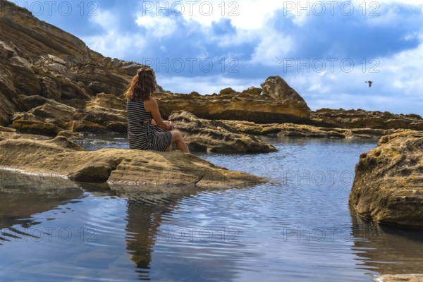 Woman sitting on ancient rocks by a tranquil tidal pool, watching the calm sea and rugged coast of mount jaizkibel in the basque country, finding solitude in nature